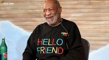 A person with their face obscured sits in front of a wooden panel wall, wearing a black sweatshirt emblazoned with “HELLO FRIEND” in rainbow colors and “FUNNY OR DIE.”